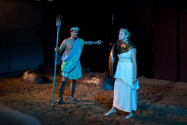 Two actors in a theatrical scene on a sandy stage. One is dressed as Poseidon with a trident, wearing a blue toga and crown. The other actor wears a white gown, gold armor, and a headpiece, holding a large shield. Rocks are scattered in the background.