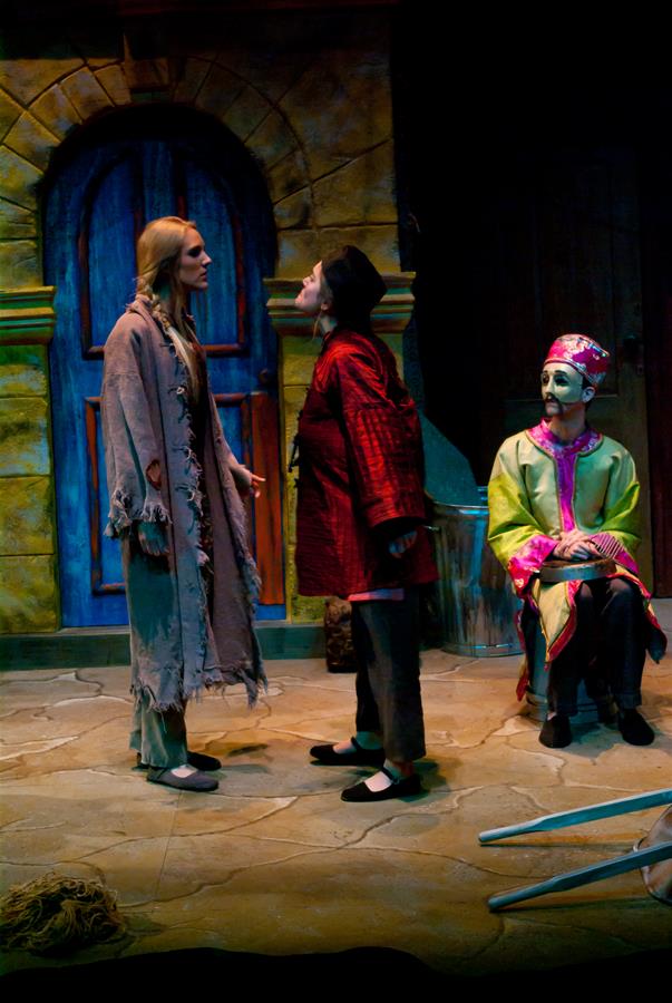 A theatrical scene with three actors: one in a tattered cloak, another in a red and black outfit, and a third seated in an ornate costume. They perform on a stone floor with arched doorways in the background.