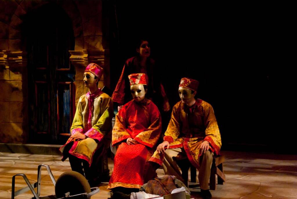 Three people in colorful, ornate costumes and masks sit on a stage with dim lighting. A fourth person stands behind them. The backdrop resembles ancient architecture, adding to the theatrical atmosphere. A prop ladder and tools are visible in the foreground.