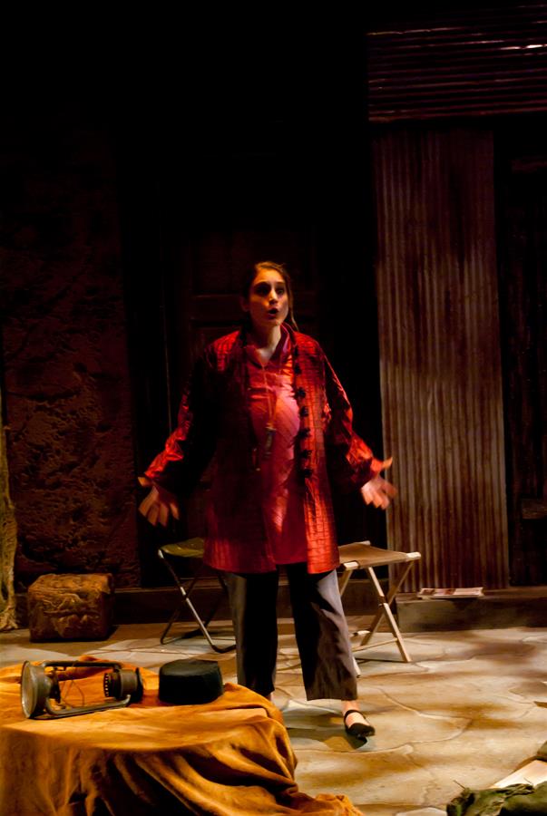 A person wearing a red top and dark pants is standing on a dimly lit stage with arms outstretched. There are chairs and a table with a cloth and objects nearby, creating a dramatic scene.