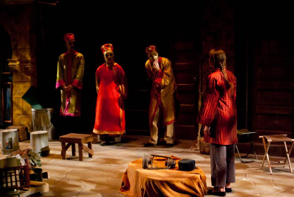 A theatrical scene with four actors on stage, three dressed in red and gold robes with hats, facing a person in a red striped garment. The stage is set with various small props and a dimly lit, rustic background.