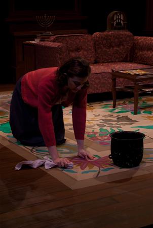 A woman wearing a red sweater and dark skirt is on her hands and knees, scrubbing a carpet with a cloth. A dark bucket is nearby. The background includes a patterned rug, a sofa, and a table with magazines.