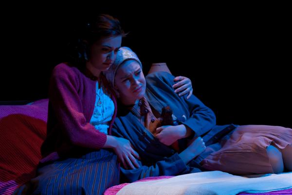 Two people sit on a bed in a dimly lit scene. One is comforting the other, who looks distressed and is holding a stuffed bear. They are dressed in vintage-style clothing, suggesting a period setting.
