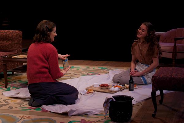 Two people sitting on a patterned floor stage set, engaging in a conversation. They are having a picnic with food and a bottle of drink on a white cloth. The background features dim lighting and vintage furniture, creating an intimate setting.