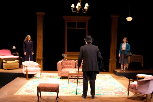 A theater scene depicting an older man with a cane facing two younger women in a living room set. The stage includes armchairs, a sofa, a chandelier, and a window frame, with warm lighting highlighting the actors.