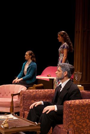 Three actors on stage perform a dramatic scene in a living room setting. A man in a suit sits on a patterned armchair. Two women, one in a floral dress standing and another in a blue jacket seated, face him. Vintage furniture decorates the stage.