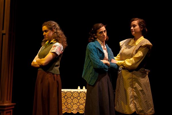 Three women stand on a dimly lit stage. The woman on the left looks away with arms crossed. The middle woman wears a blue cardigan and stands facing the camera. The third woman, in a patterned dress, stands close to the middle woman with arms linked.