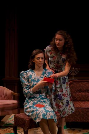 Two women in floral dresses are in a dimly lit room. One is seated, holding a red object, while the other stands beside her, leaning in. A menorah is visible in the background on a brown couch.