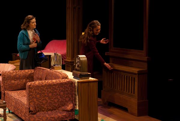 Two women are on stage in a theatrical setting. One woman, wearing a blue sweater, stands watching the other, who is wearing a dark red dress and interacting with a wooden furniture piece. The scene suggests a domestic interior with a couch and other furnishings.