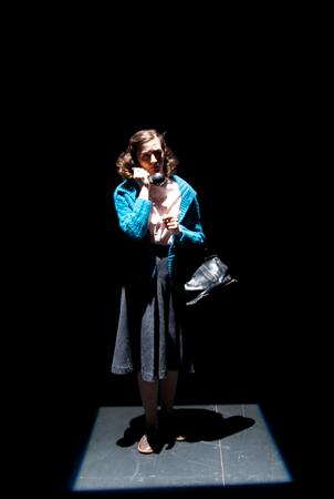A person stands alone in a spotlight on a dark stage, wearing a teal cardigan, white blouse, and gray skirt. They hold a phone to their ear and have a black bag. The scene is dramatic and emphasizes solitude.