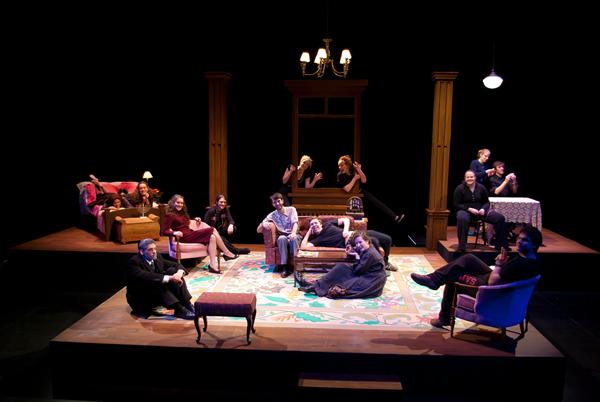 A group of people is sitting and interacting on a stage set designed like a living room, featuring a colorful rug, furniture, and a chandelier. The lighting is dim, creating an intimate atmosphere.