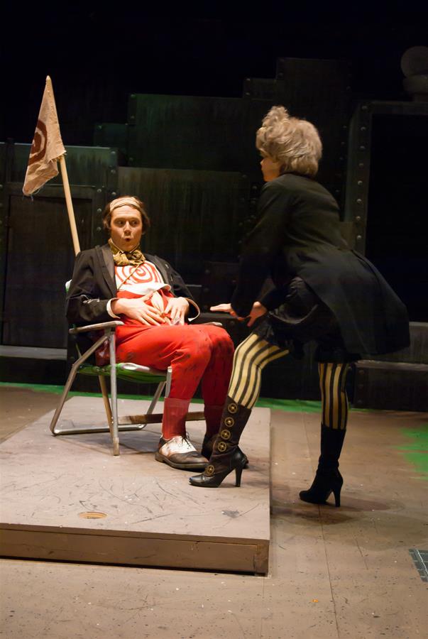 A person in a chair with vibrant red pants and a headband sits on stage. Another person faces them, wearing a black outfit with striped leggings and high-heeled boots. The set features a dark backdrop with industrial elements and a small flag.