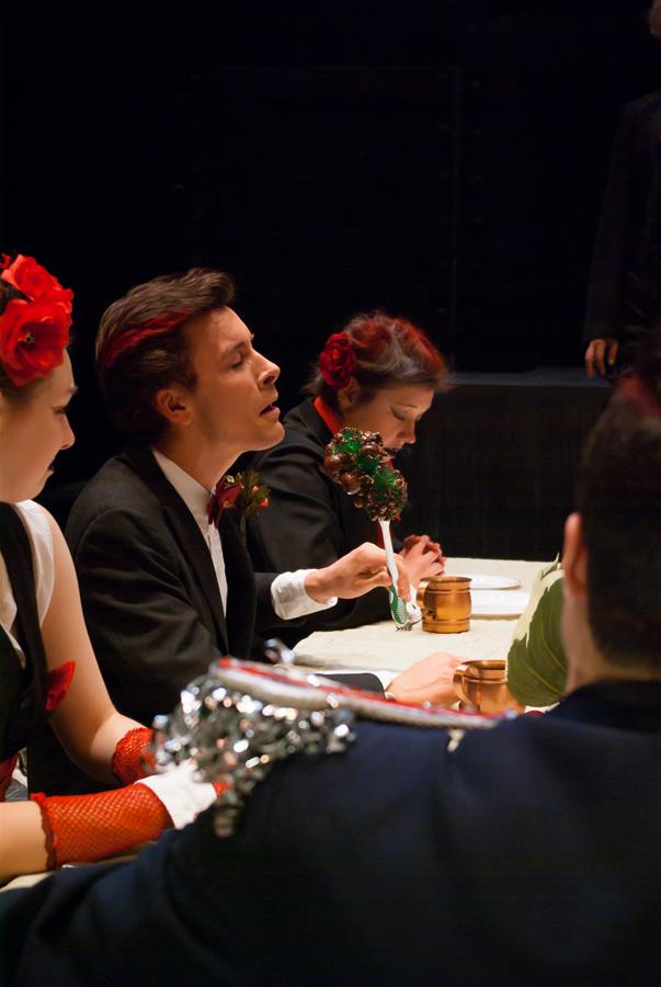 People are seated at a table, dressed in formal attire with red accents. One person appears to be speaking, holding a decorative object, while others listen intently. The mood is theatrical and lively.