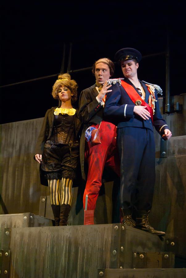 Three actors in theatrical costumes stand on stage. The left actor wears striped pants and a black jacket, the middle actor wears a red suit, and the right actor wears a navy uniform with a hat, posing dramatically against a dark backdrop.