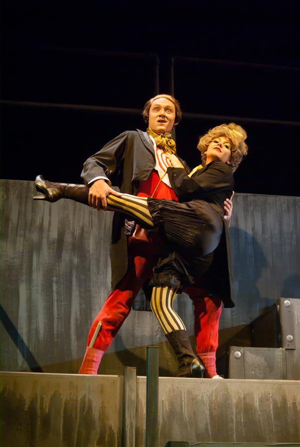 Two actors perform on stage. One wears a red and black costume with yellow stripes and lifts the other, who is dressed in a black outfit with a short skirt and high-heeled boots. The background is a simple, industrial-style set.