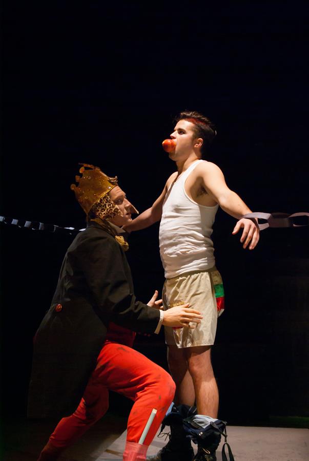 A man in a crown and red pants kneels in front of another man wearing a white tank top and shorts, who has an apple in his mouth. The scene appears theatrical, with dramatic lighting and costumes.