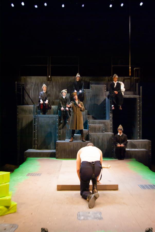 A person kneels on stage, facing away, while five others in dark clothing and hats watch from elevated steps, one in a brown coat holding a microphone. The stage is set with a multi-level industrial backdrop under dim lighting.