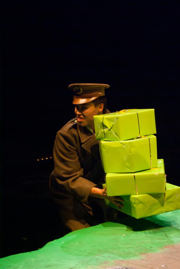 A person in a military uniform balances a stack of three bright green gift boxes. The background is dark, highlighting the focus on the wrapped gifts and uniformed individual.