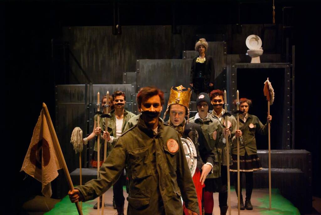 Six performers on stage in theatrical costumes and props, including military uniforms and medieval attire. One holds a flag, and others have various accessories. The set features stairs and a raised platform with a throne-like chair in the background.