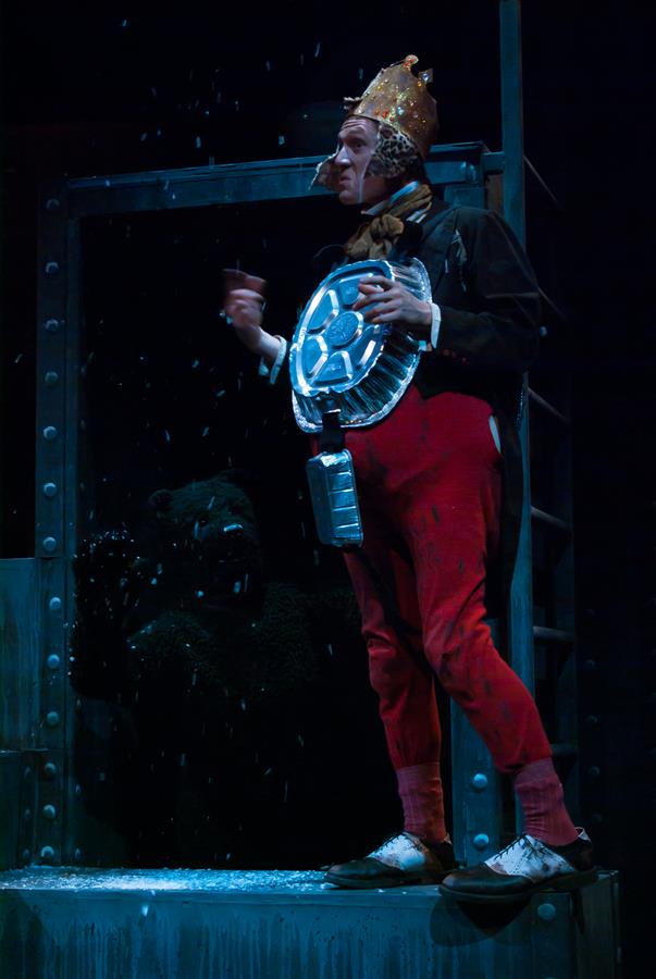 A person dressed as a soldier with a bucket helmet and holding a trash can lid stands on stage. They are wearing red pants and standing in front of a bear costume in a dark, snowy setting.