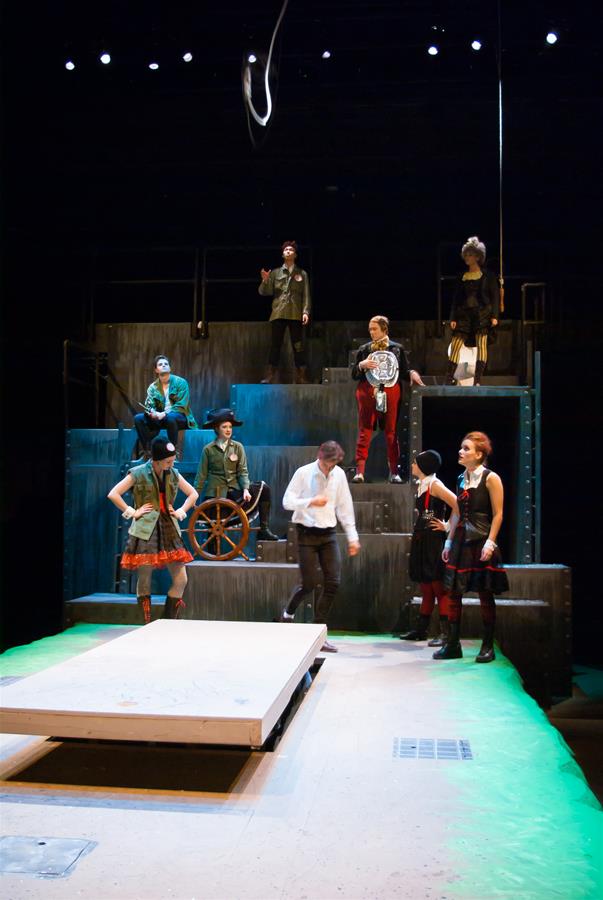 A group of people, dressed in eclectic costumes, stand and sit on a multi-level stage set. One person holds a ship's wheel, and others sport various outfits. The scene is lit with a spotlight against a dark backdrop, creating a theatrical atmosphere.