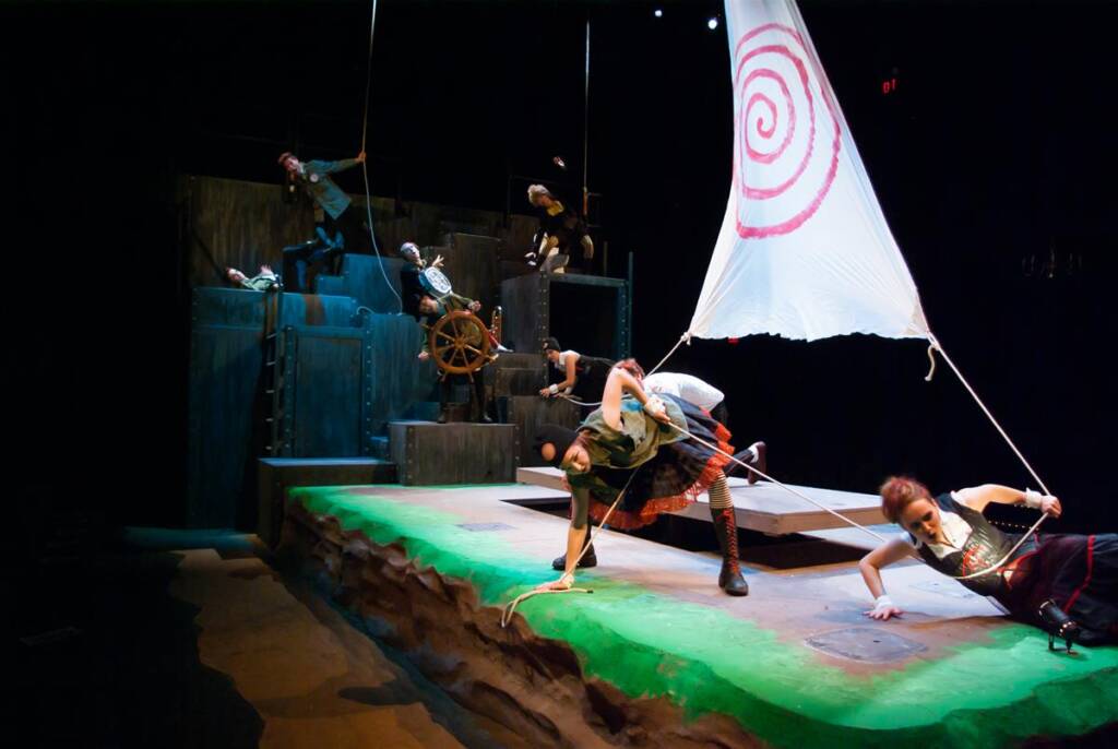 Performers on stage act in a dynamic scene with a large white sail marked with a red spiral. The set resembles a ship with ropes and a steering wheel. The scene is lively, with actors in various poses suggesting movement and action.