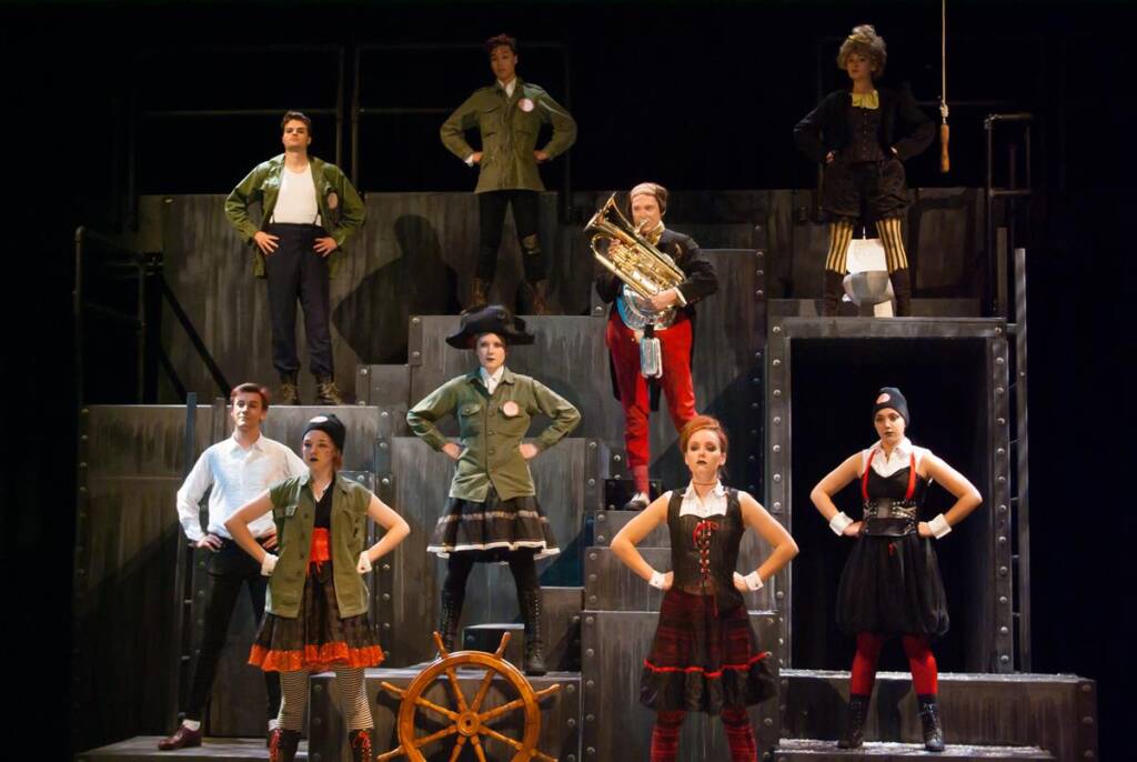 Eight performers in theatrical costumes stand on a multi-level stage. One holds a large brass instrument, while another stands by a ship's wheel. The scene is dramatic, with bold outfits and confident poses against a dark backdrop.