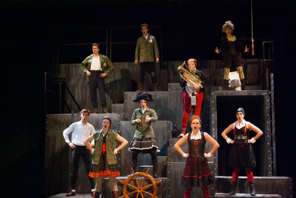 A group of performers on stage in colorful costumes. They stand on tiered platforms with a person in the center holding a tuba. A wooden steering wheel is in front, and the backdrop is dark, creating a dramatic setting.
