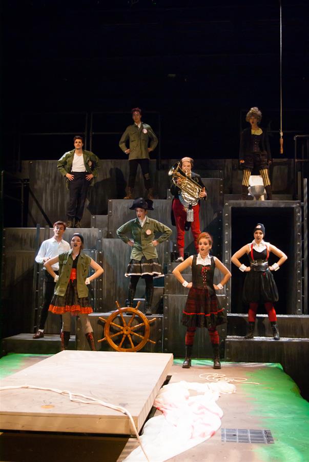 A group of performers stands on a tiered stage set resembling a ship, with metal railings and a wooden steering wheel. Some hold instruments and wear costumes suggesting a nautical theme. The stage is lit, highlighting the ensemble.