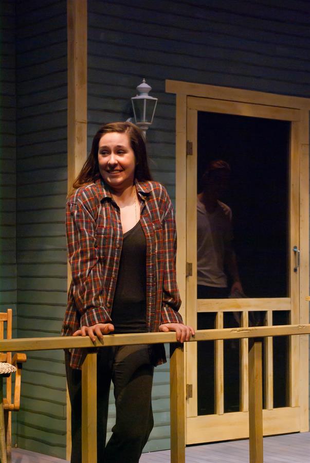 A woman in a plaid shirt stands on a porch, leaning on the railing with a smile. A dimly lit figure is behind the screen door. The scene suggests a casual, evening setting.