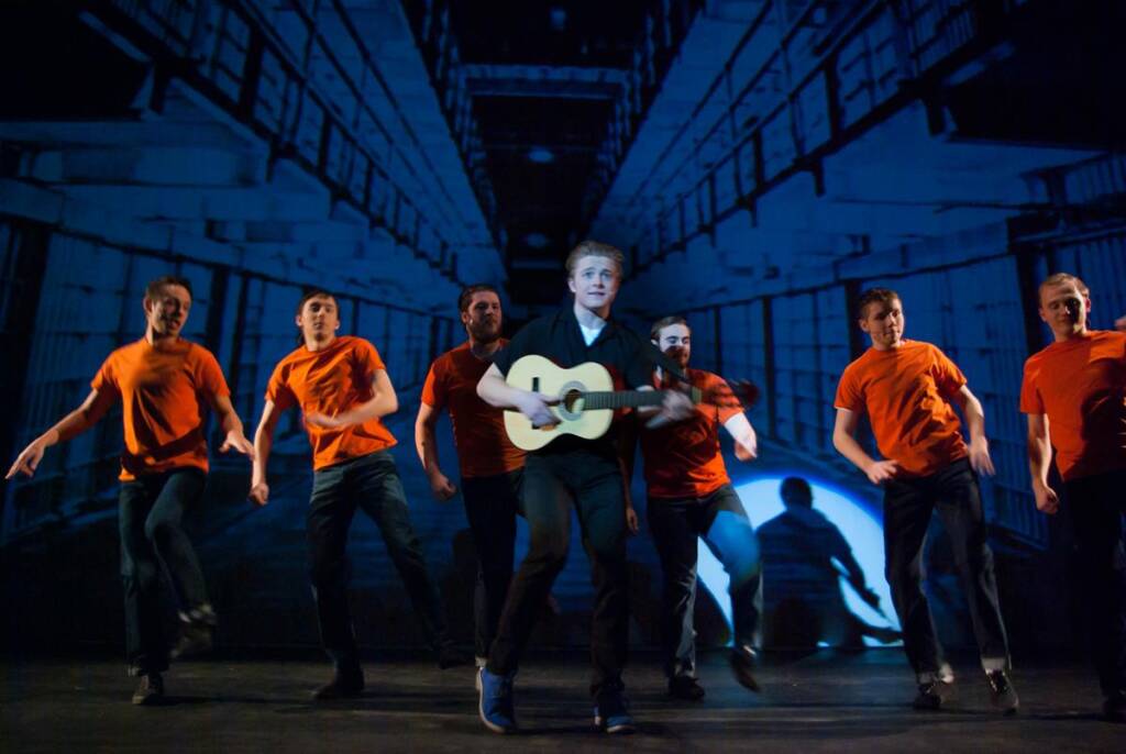 A group of seven people performing on stage. One person in the center is playing a guitar, surrounded by others in orange shirts and jeans, dancing energetically. The background displays an image resembling a prison hallway.