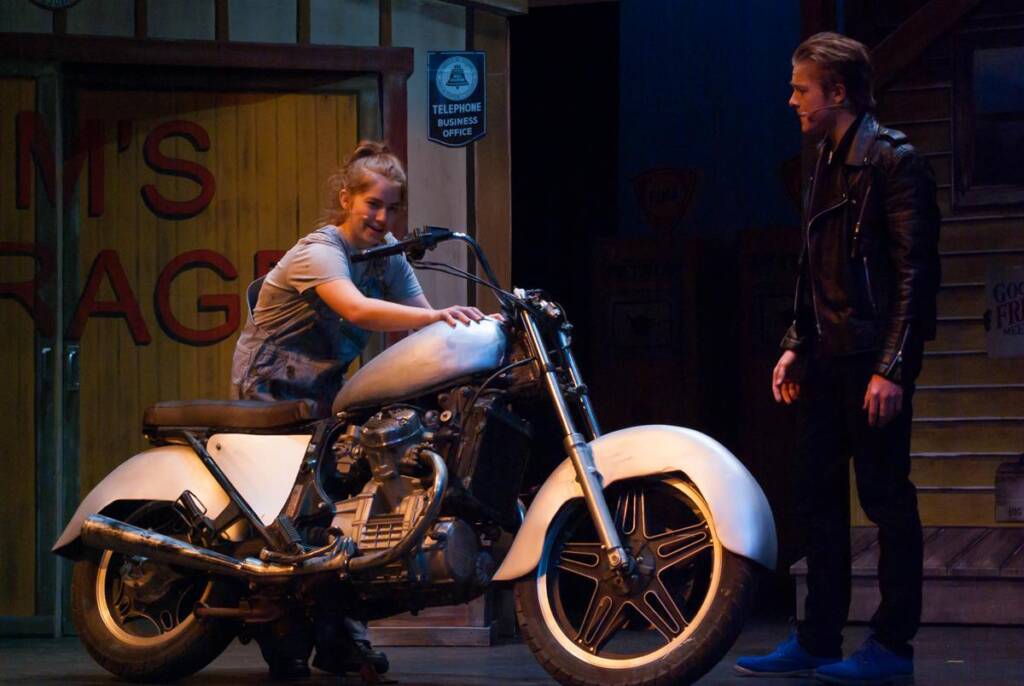 A young person sits on a white motorcycle, smiling, while another person in a black leather jacket stands nearby watching. They are on a stage with a wooden garage backdrop and vintage telephone booth sign.