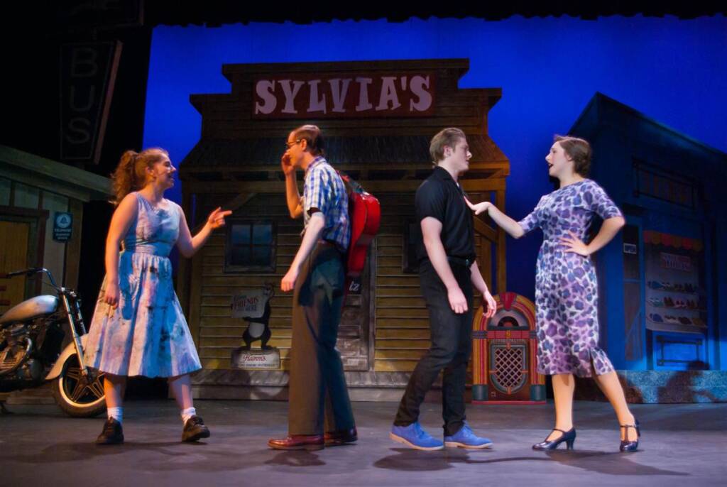 Four actors perform on stage in front of a set resembling an old storefront labeled "Sylvia's." One actor in a blue dress gestures animatedly, while others stand in varied poses. A motorcycle and vibrant backdrop enhance the scene.