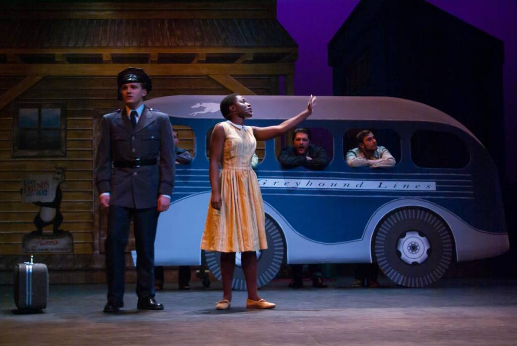 A theatrical scene with a woman in a yellow dress gesturing in front of a vintage Greyhound bus and a man in a uniform standing beside her. Three people are seated inside the bus, and a station is in the background.