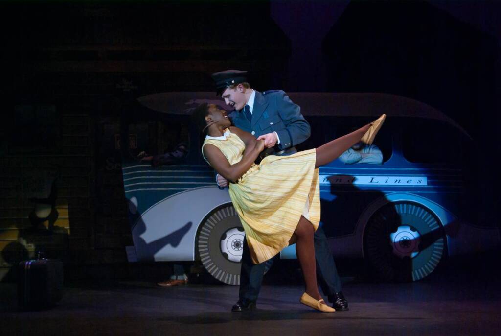 A woman in a yellow dress and a man in a blue uniform dance dramatically on stage, with the woman leaning back and lifting a leg. Behind them, a vintage bus and suitcase are visible, creating a theatrical and nostalgic scene.