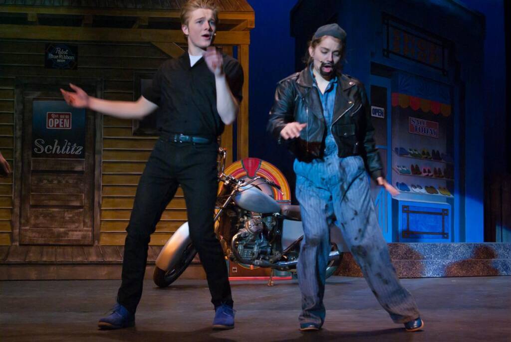 Two performers are on stage in front of a wooden building and a motorcycle. One wears a black outfit, and the other is dressed as a biker with a fake beard, leather jacket, and cap. Both seem to be in motion, possibly dancing or acting.