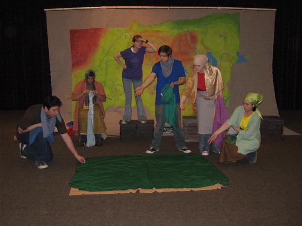 Six people are performing on a stage with a colorful backdrop. They are dressed in casual and costume-like clothing, interacting with a green fabric laid out on the floor. The scene suggests a theatrical play or rehearsal.