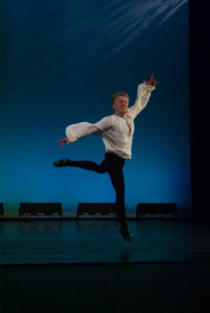 A dancer in a white shirt and black pants performs a graceful leap on stage. The background is a gradient of blue, enhancing the elegance of the performance. The dancer's expression is focused and joyful.