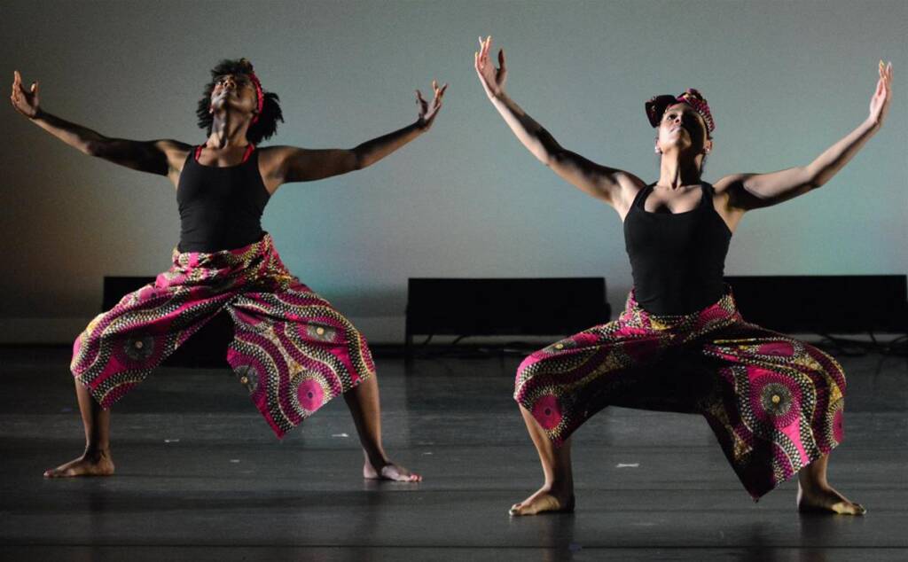 Two dancers perform on stage, wearing patterned pants and headbands. They have their arms raised, with legs bent in a wide stance. The background is a plain, softly lit wall.