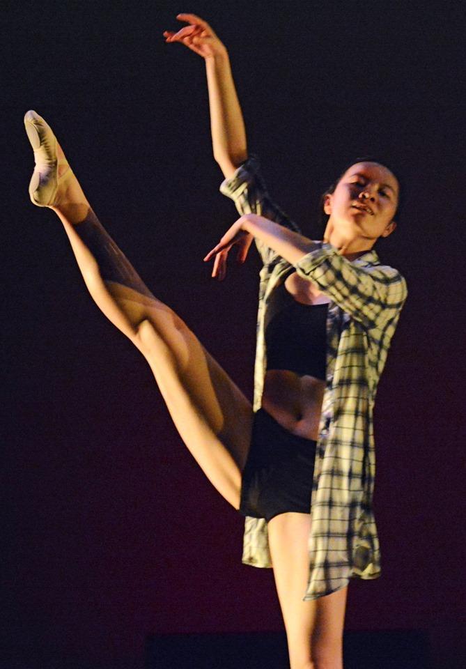 A dancer gracefully balances on one foot, wearing a plaid shirt over a black top and shorts. Her extended leg is raised high, and her arms are elegantly curved. The background is dark, highlighting her poised movement.
