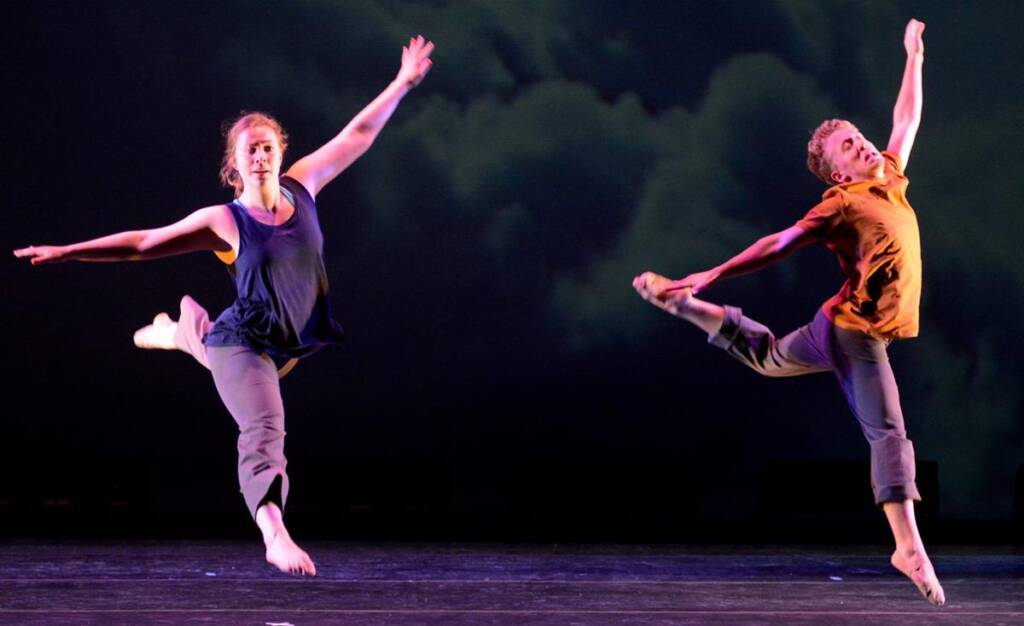 Two dancers are mid-air, performing a leap on stage. The one on the left wears a dark tank top and gray pants, and the one on the right wears a brown shirt and gray pants. The background is a dark, cloudy sky.