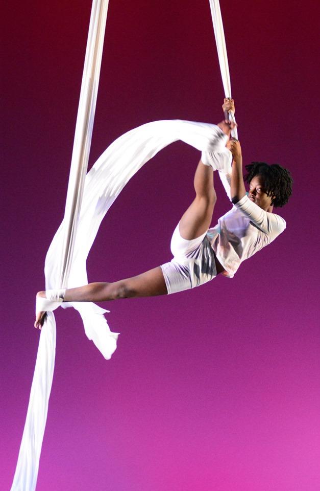 A person performing an aerial silk routine, suspended mid-air against a gradient purple background. They are wrapped in white silks, holding onto them with one hand and one foot, while extending the opposite leg gracefully.