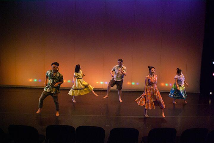 Five dancers perform on a stage with colorful lighting. They are wearing bright, patterned outfits and are mid-motion, each in a different pose. The backdrop is a simple, illuminated wall. The audience seating is visible in the foreground.