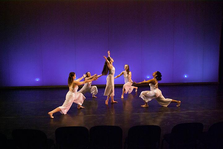 Five dancers wearing white outfits perform gracefully on stage, surrounded by a purple-lit backdrop. They are arranged in a circular formation, with one dancer in the center extending her arms upward.