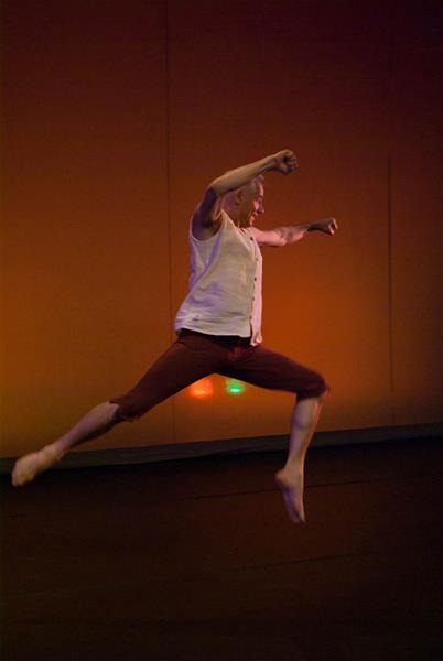 A dancer performs a mid-air leap on stage, wearing a white sleeveless top and brown pants. The background is a warm, softly lit hue, creating a dynamic and expressive atmosphere.