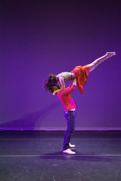 A dancer in a red jacket lifts another dancer in a red skirt and striped top, creating a dynamic pose against a purple-lit stage background.