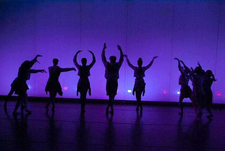 Silhouettes of a group of dancers in expressive poses against a background lit with purple and blue lights.