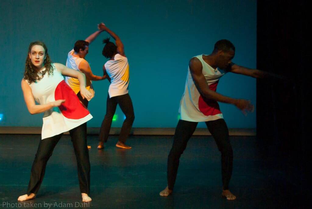 Four dancers perform on stage, wearing white tunics with red and yellow patterns. Two are in the foreground, one with arms crossed and the other with arms extended. Two dancers in the background are mid-movement, with arms raised.