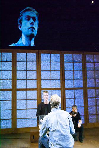 A large screen displays a man's face above a stage where two people are seated on chairs facing each other. The backdrop features a wall with a grid pattern resembling shoji screens. A third person crouches next to the seated people.