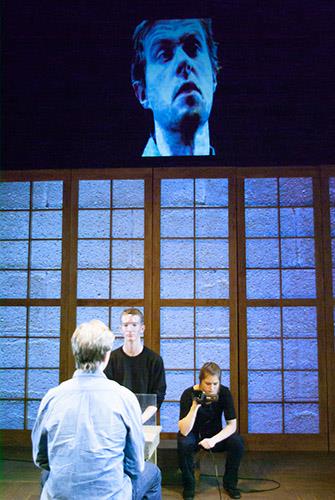 A stage scene with three individuals indoors. One person sits with their back facing the camera, another sits on the floor with a phone, and the third stands near. A large, projected face is displayed above them on a screen.
